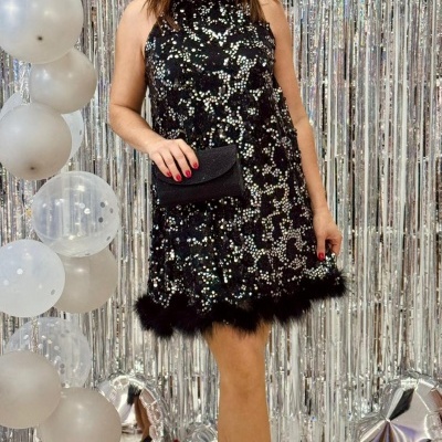 Mulher com vestido preto de lantejoulas e penas, carteira preta e sapatos pretos em ambiente festivo com balões prateados.