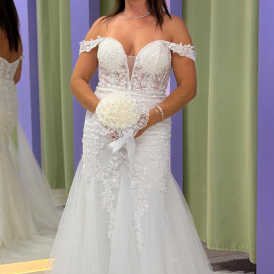 Mulher a usar vestido de noiva branco com detalhes de renda e tule, segurando bouquet de rosas brancas, junto a fundo com arcos lilás e cortinas verde-claras.