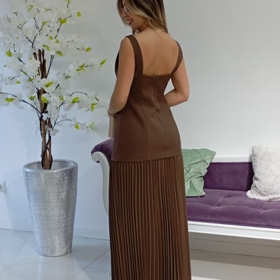 Vestido castanho com saia plissada, visto por trás, em ambiente interior com vaso prateado e sofá roxo