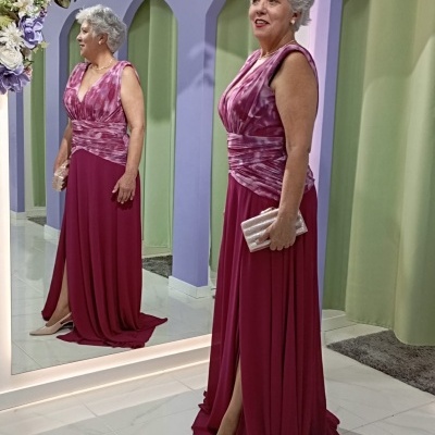 Mulher com vestido longo vinho e rosa em frente a espelho com decoração floral e cortinas verdes.