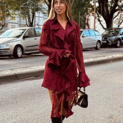 Mulher com vestido vermelho vinho com folhos e renda, botas pretas e mala preta numa rua com árvores e carros.