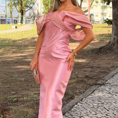 Mulher em vestido longo cor-de-rosa acetinado com folhos nos ombros, usando óculos, carteira brilhante e sandálias brancas num parque