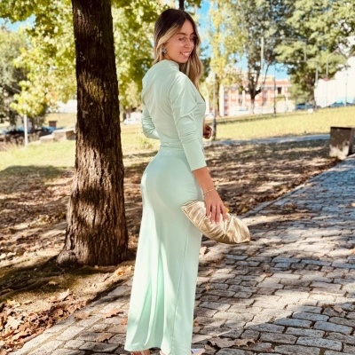 Mulher com vestido verde longo e bolsa dourada em passeio de calçada com árvores