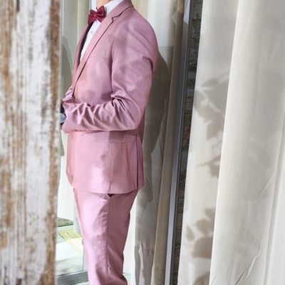 Jovem com fato rosa e gravata borboleta posando junto a cortinas bege