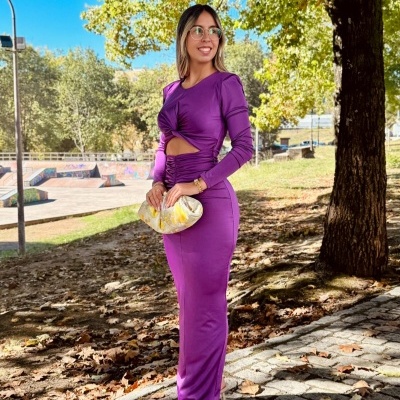 Mulher a usar vestido lilás com recorte e mala clutch creme num parque ao ar livre.