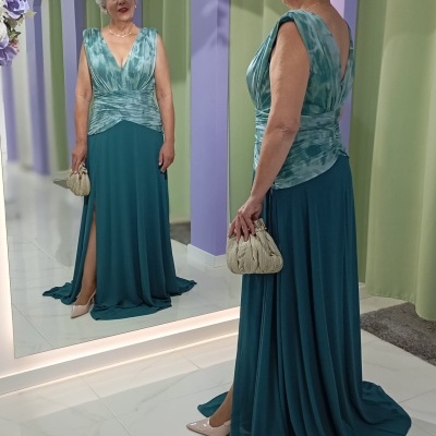Mulher com vestido comprido azul esverdeado e parte superior azul claro franzida, segurando mala bege, de frente para espelho num espaço com paredes lilases e arco de flores