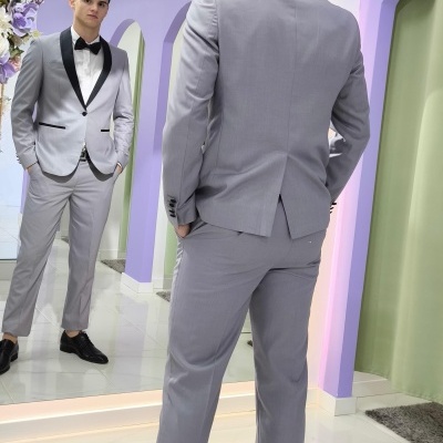 Homem a usar fato cinzento claro com gravata borboleta preta perante espelho em ambiente moderno com decoração floral.