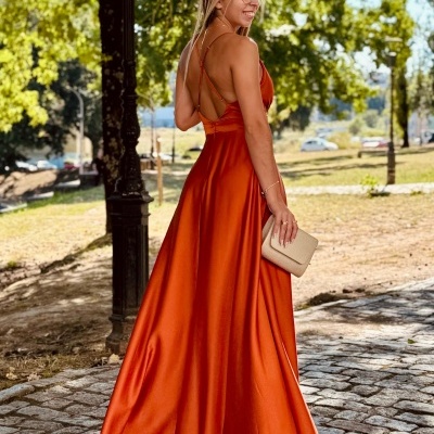 Vestido longo laranja de cetim com alças cruzadas e carteira bege numa paisagem ao ar livre