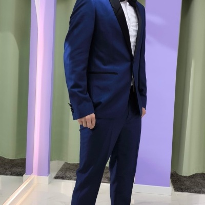 Homem de terno azul com lapelas pretas e gravata borboleta preta em interior com parede lilás