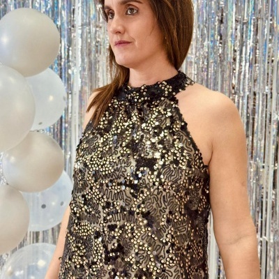 Vestido preto com lantejoulas douradas e prateadas, balões brancos e cortina prateada ao fundo.