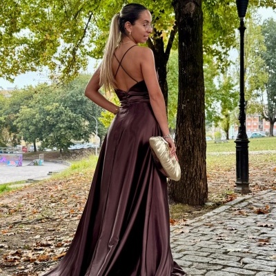 Mulher de costas com vestido longo castanho de cetim e mala dourada ao ar livre