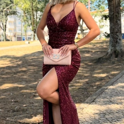 Mulher com vestido longo borgonha brilhante e clutch rosa num parque