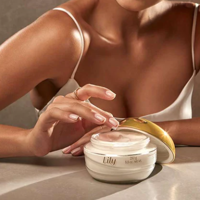 Frasco de creme corporal Lily com tampa dourada e mão feminina a tocar o creme