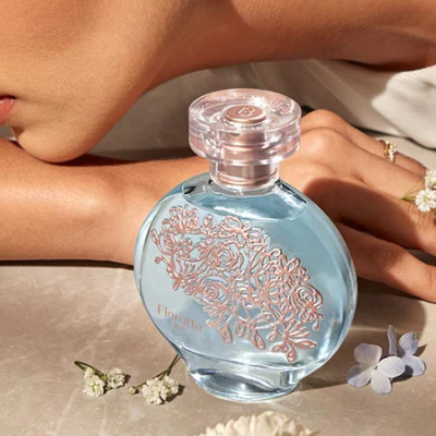 Garrafa de perfume azul claro com detalhes florais rosas e tampo transparente, junto a flores pequenas numa superfície clara