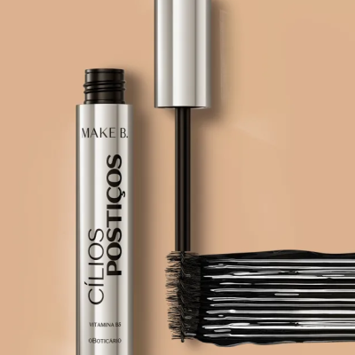 Máscara de pestanas preta MAKE B. em embalagem prateada com pincel ao lado, fundo bege