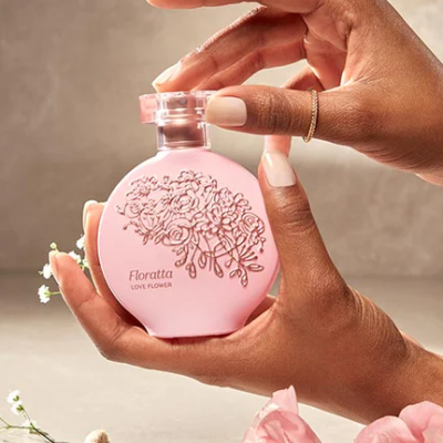 Frasco de perfume rosa claro com relevo floral e texto, segurado por mãos femininas com unhas manicadas.