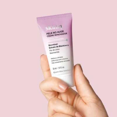 Tubo pequeno de creme renovador rosa com texto em preto e roxo, segurado por uma mão sobre fundo rosa claro.