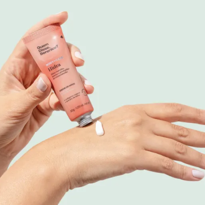 Mãos aplicando creme Hidra, tubo cor de salmão, fundo cinza