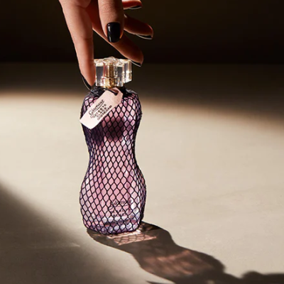 Frasco de perfume feminino rosa com rede preta e etiqueta branca sendo segurado por uma mão com unhas pintadas de negro