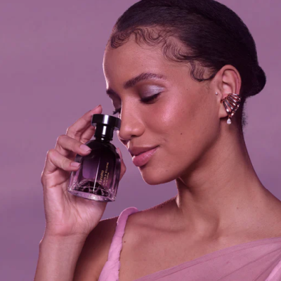 Mulher segurando frasco de perfume preto com líquido translúcido em fundo rosa lilás