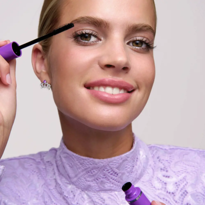 Mulher a aplicar máscara de pestanas com aplicador preto e cabo roxo, usando blusa lilás de renda.