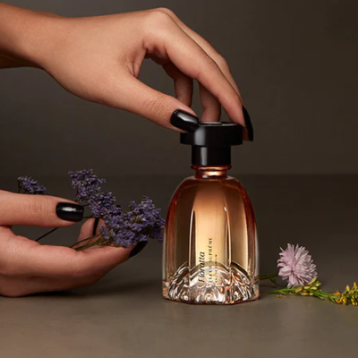 Frasco de perfume âmbar com tampa preta e base de vidro trabalhado, mãos femininas segurando e flores ao redor