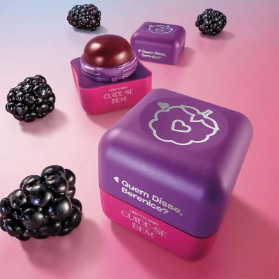 Produto cosmético com embalagem rosa e roxa ao lado de amoras pretas sobre fundo rosa.