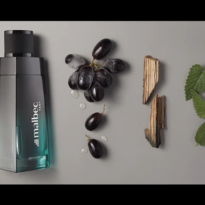 Frasco de perfume Malbec O Boticário com uvas, gotas, madeira e folhas sobre fundo cinza.