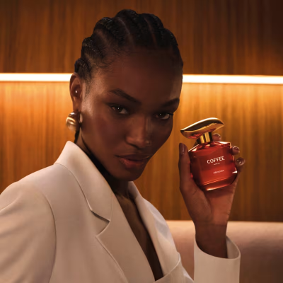 Mulher segura perfume vermelho COFFEE O BOTICÁRIO com fundo de madeira iluminado