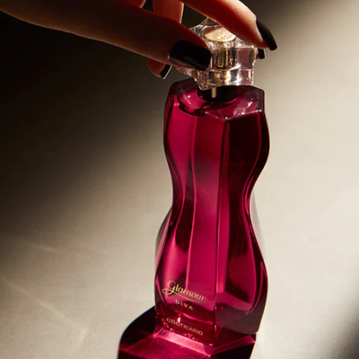 Frasco de perfume rosa escuro com tampa transparente e texto dourado, mão segurando a tampa.