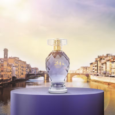 Frasco de perfume lilás com tampa transparente e detalhes dourados sobre pedestal roxo com vista de rio e edifícios históricos ao fundo
