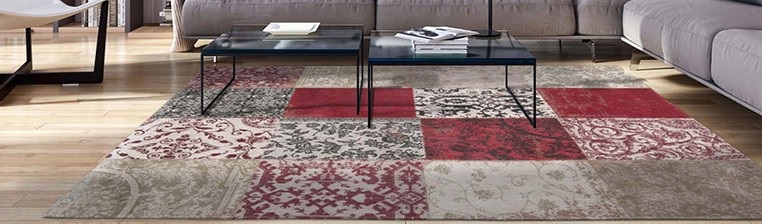 Tapete decorativo com padrão quadrado em tons de bege, vermelho e bordô numa sala com sofá cinza.