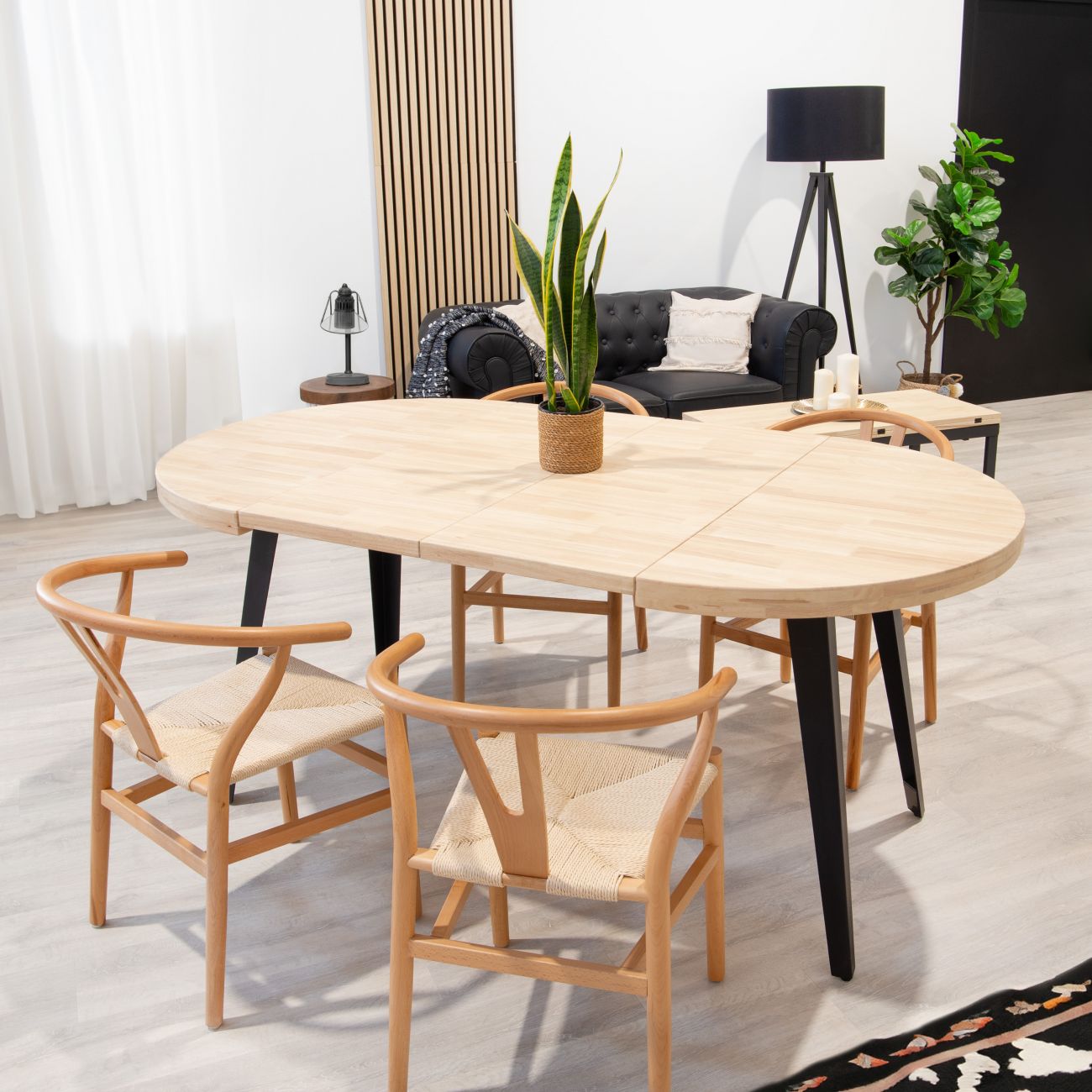 Mesa de madeira clara com cadeiras, planta e sofá num ambiente interior moderno