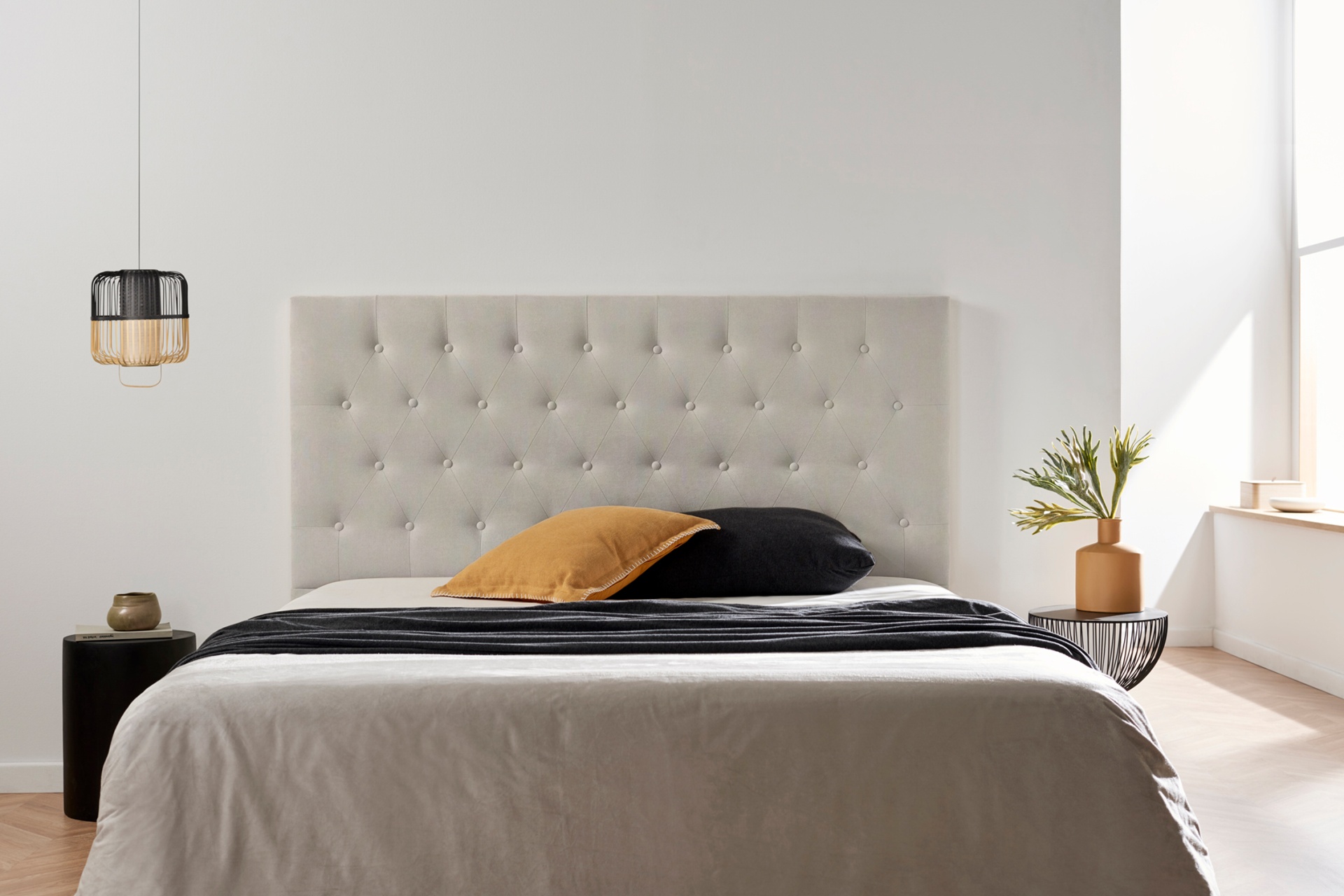 Cama com cabeceira estofada bege e roupa de cama em tons cinza e preto, com almofadas preta e castanha, junto a mesa lateral preta com vaso castanho e planta, candeeiro pendente preto e parede branca.