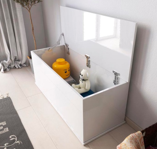 Caixa de armazenamento branca aberta com brinquedos, situada num chão claro junto a uma parede e cortina cinzenta