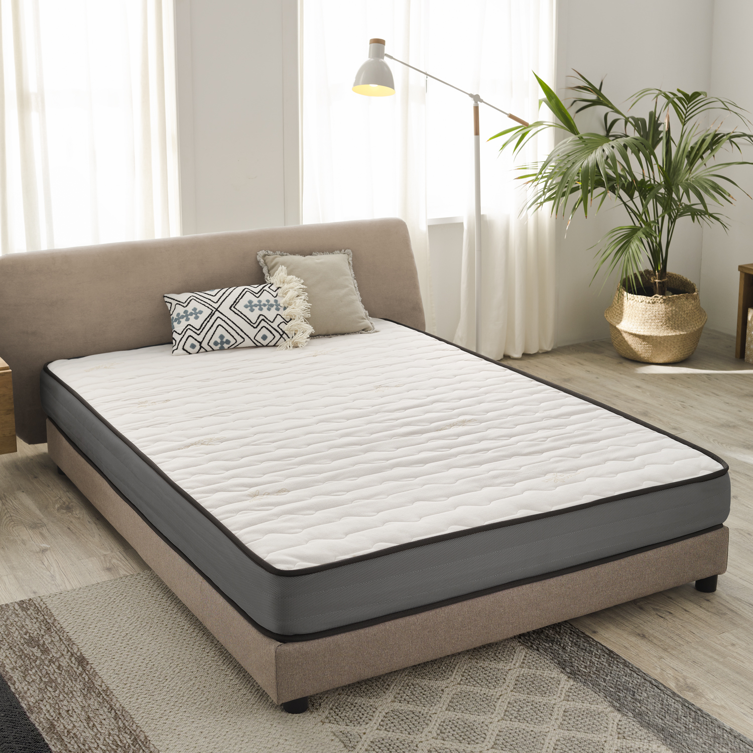 Cama estofada bege com colchão branco e almofadas decorativas, candeeiro branco e planta em vaso dourado