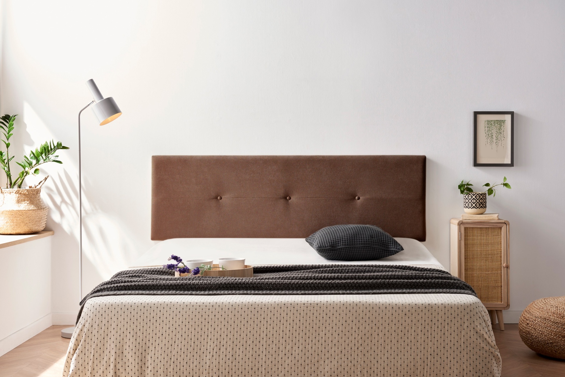 Quarto com cama de casal, cabeceira castanha, roupa de cama bege e cinza, candeeiro branco, mesa de vime com planta e quadro na parede