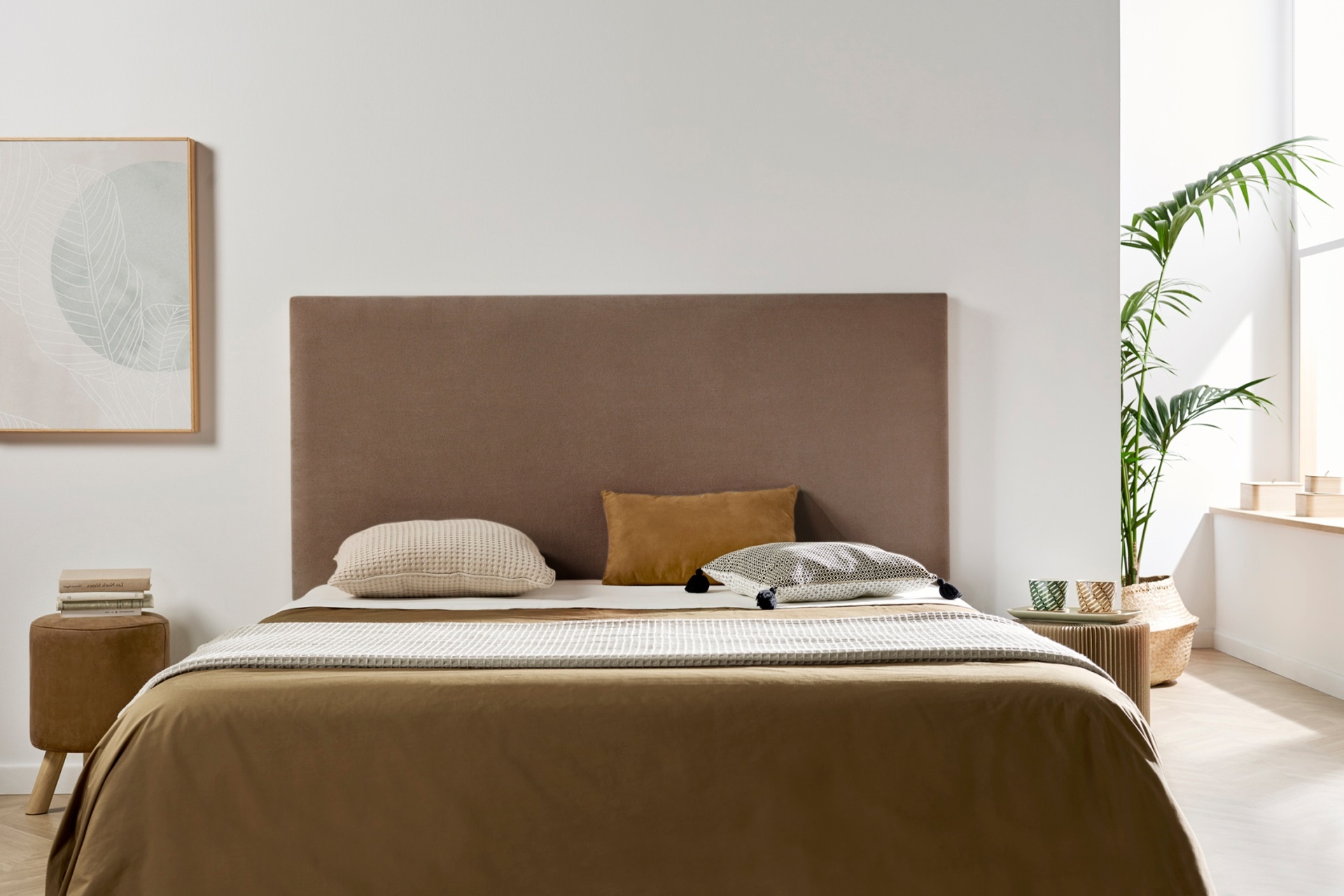 Cama com cabeceira castanha e roupa de cama castanha e cinza, mesas laterais com objetos decorativos em quarto luminoso.