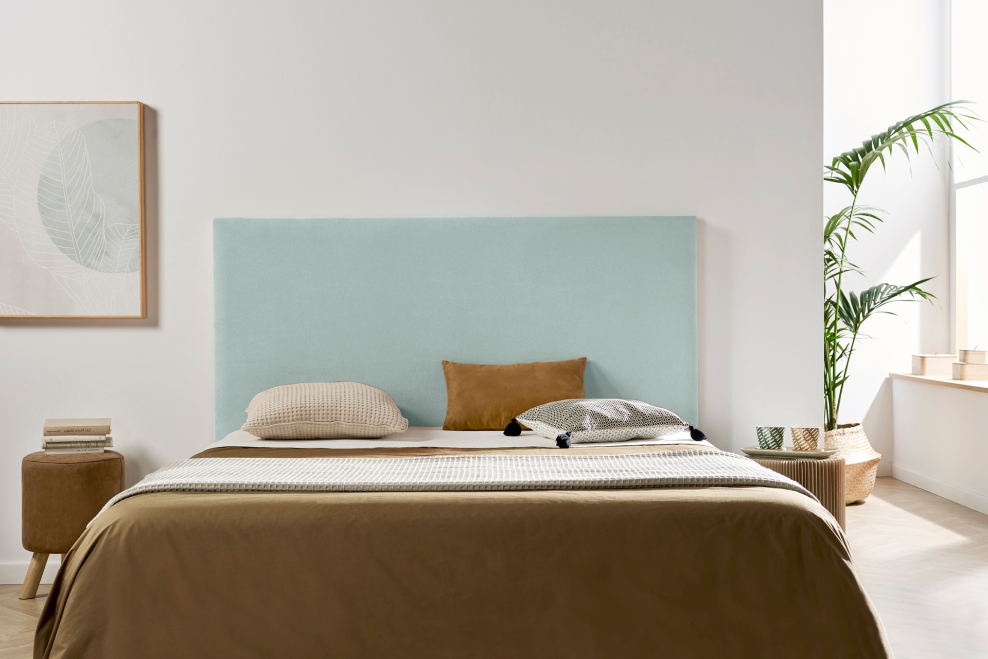 Cama com cabeceira azul clara, roupa de cama castanha e bege, mesa de cabeceira com livros e planta junto a janela