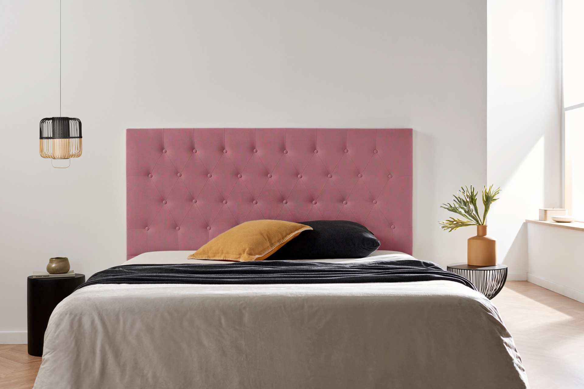 Cama com cabeceira rosa estofada, roupa de cama cinza, almofadas preta e mostarda, candeeiro pendurado e vaso com plantas