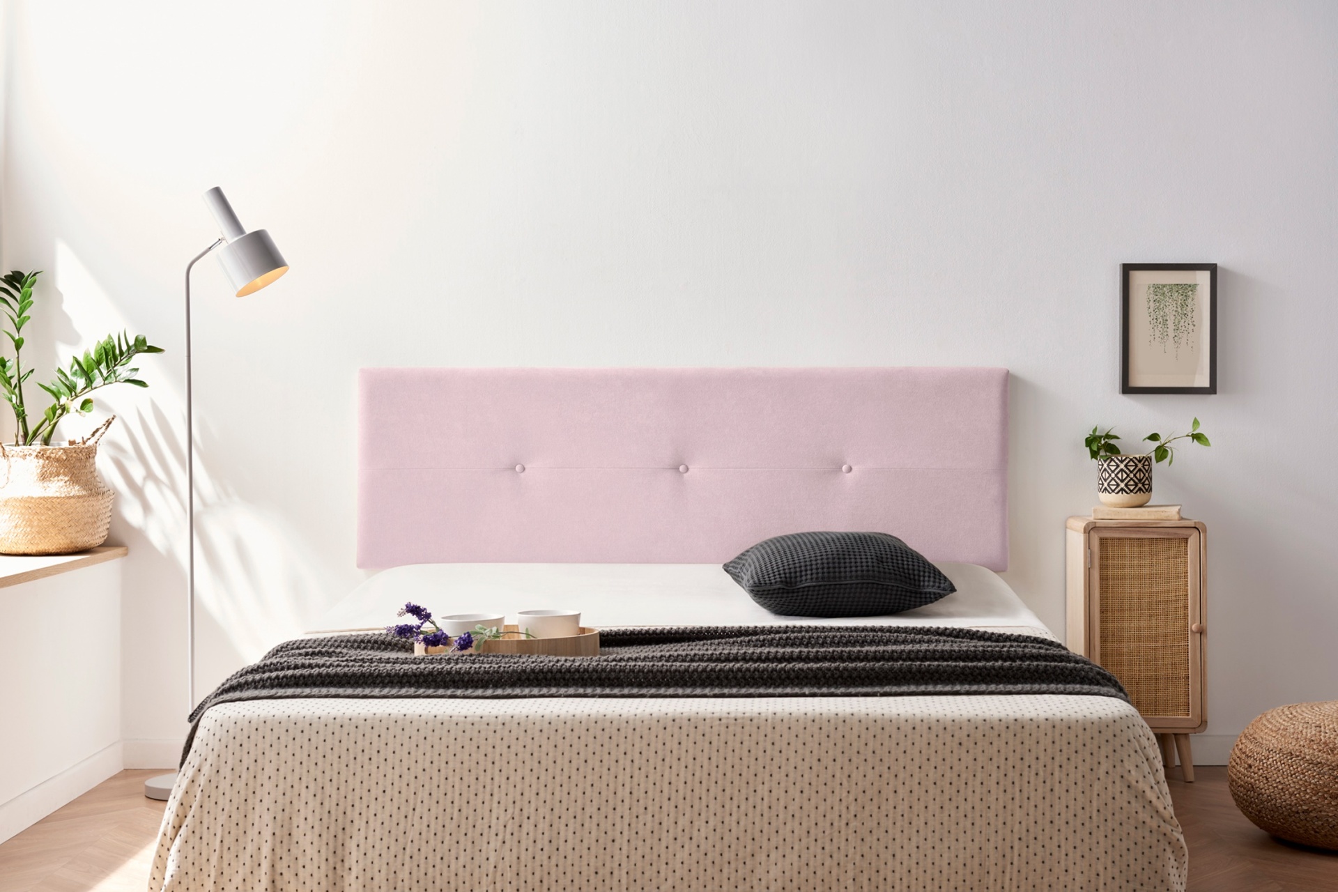 Quarto com cama de cabeceira rosa, roupa de cama bege com padrão, candeeiro, aparador e planta