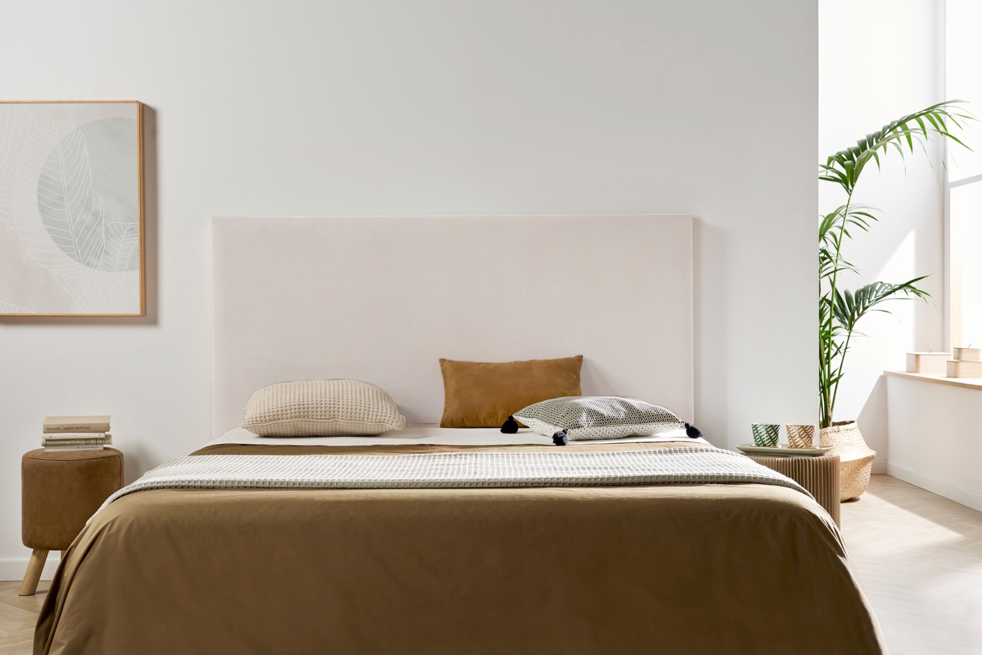 Quarto com cama de tons castanho e bege, mesa de cabeceira em madeira com livros, quadro decorativo branco e planta verde junto à janela