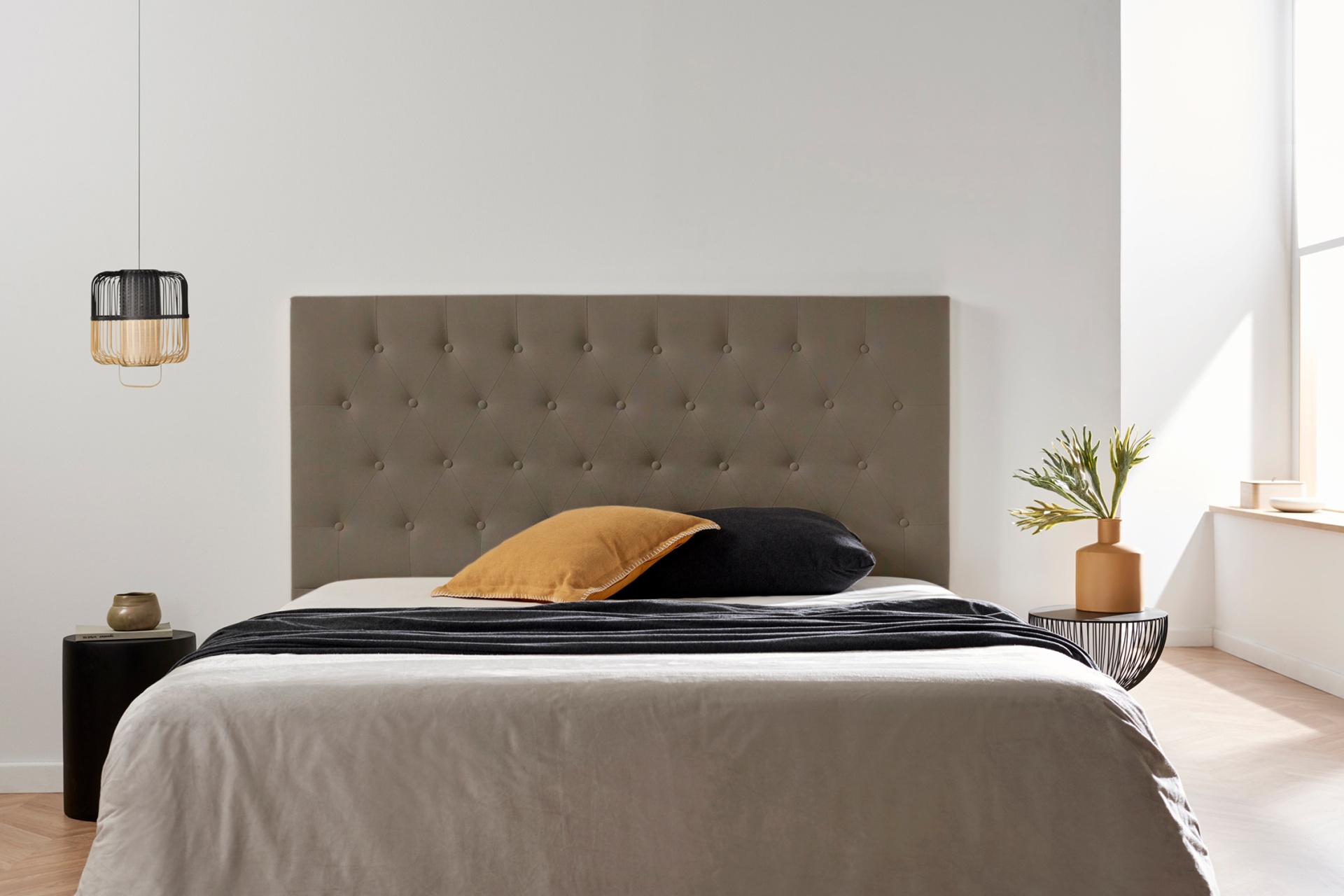 Cama com cabeceira estofada cinza, roupa de cama cinza e preta, almofadas bege e preta, luminária pendente, mesa lateral com vaso e planta