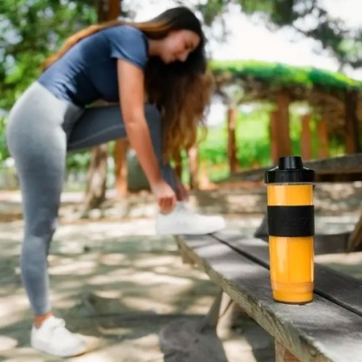 Garrafa térmica laranja com tampa preta numa bancada de madeira ao ar livre, mulher a amarrar sapatos ao fundo
