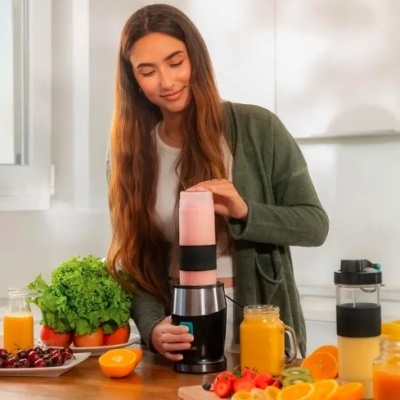 Liquidificador portátil com batido rosa e mulher a usar na cozinha com frutas e legumes