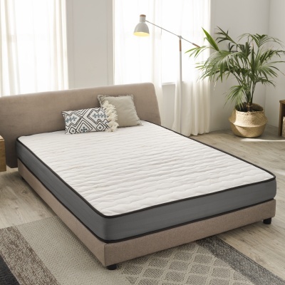 Cama estofada bege com colchão branco e almofadas decorativas, candeeiro branco e planta em vaso dourado