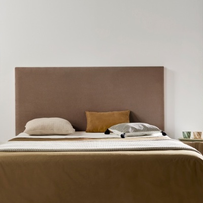 Cama com cabeceira castanha e roupa de cama castanha e cinza, mesas laterais com objetos decorativos em quarto luminoso.