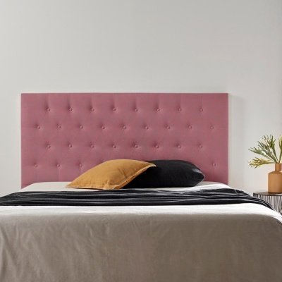 Cama com cabeceira rosa estofada, roupa de cama cinza, almofadas preta e mostarda, candeeiro pendurado e vaso com plantas