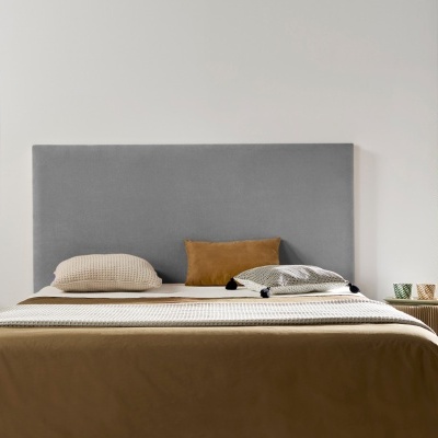 Cama com cabeceira cinza e roupa de cama castanha, almofadas variadas, mesa de cabeceira e planta verde