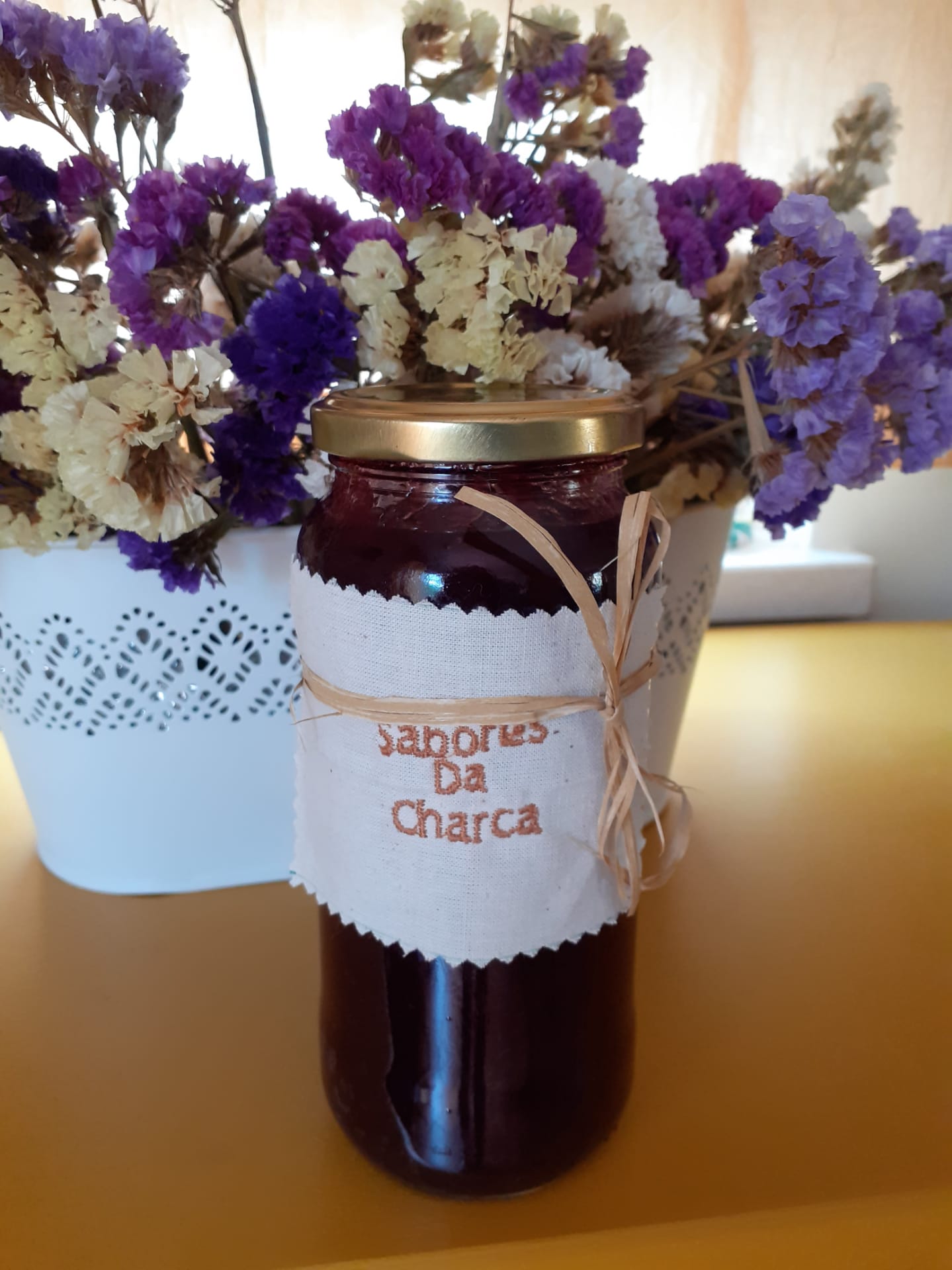 Garrafa de vidro com geleia vermelha e etiqueta de tecido com texto 'Sabores Da Charca' ao fundo flores secas em vaso branco.